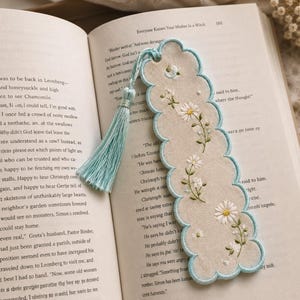 May include: A scalloped-edge fabric bookmark in a light beige color featuring embroidered white daisy flowers with yellow centers and green stems. The bookmark is finished with a light blue stitched border and a matching light blue tassel.