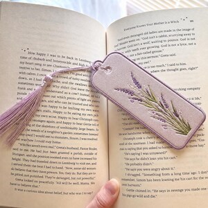 May include: A rectangular light purple fabric bookmark featuring an embroidered lavender sprig design in shades of purple and green. The bookmark includes a matching light purple tassel attached through a reinforced eyelet at the top edge.
