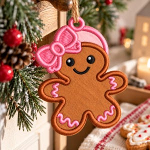 May include: A brown gingerbread person ornament featuring a pink embroidered bow on its head and pink wavy icing details on its arms and legs. The ornament has black embroidered eyes and a smiling mouth, with a loop at the top for hanging.