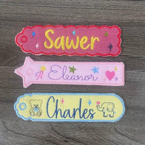 May include: Three rectangular fabric tags with embroidered names. The top tag is pink with yellow text reading Sawer and colorful stars. The middle tag is light pink with purple text reading Eleanor, stars, and a heart. The bottom tag is yellow with blue text reading Charles, a bear, an elephant, and stars.