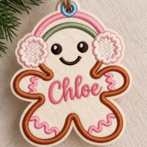 May include: A cream-colored felt gingerbread person ornament featuring embroidered brown outlines, pink wavy accents, and a smiling face. The figure wears pink and green earmuffs and has the name Chloe embroidered in pink script across the center.