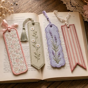 May include: Four fabric bookmarks resting on an open book. The first is floral with a pink ribbon, the second is sage green with embroidered daisies and a tassel, the third is lavender with embroidered sprigs, and the fourth has pink stripes.