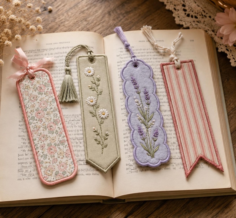 May include: Four fabric bookmarks resting on an open book. The first is floral with a pink ribbon, the second is sage green with embroidered daisies and a tassel, the third is lavender with embroidered sprigs, and the fourth has pink stripes.