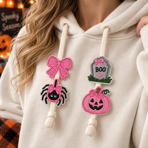 May include: A cream-colored hoodie featuring two thick drawstrings adorned with embroidered patches. The left string has a pink bow and a black spider with a pink bow. The right string has a gray tombstone with the text BOO and a pink pumpkin.