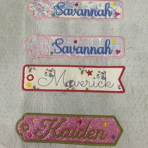 May include: Four embroidered fabric name tags. Two rectangular tags feature the name Savannah in blue script with a flower icon. One rectangular tag with a pointed end features the name Maverick in black script with floral and star accents. One pink glitter tag features the name Kaiden in green script.