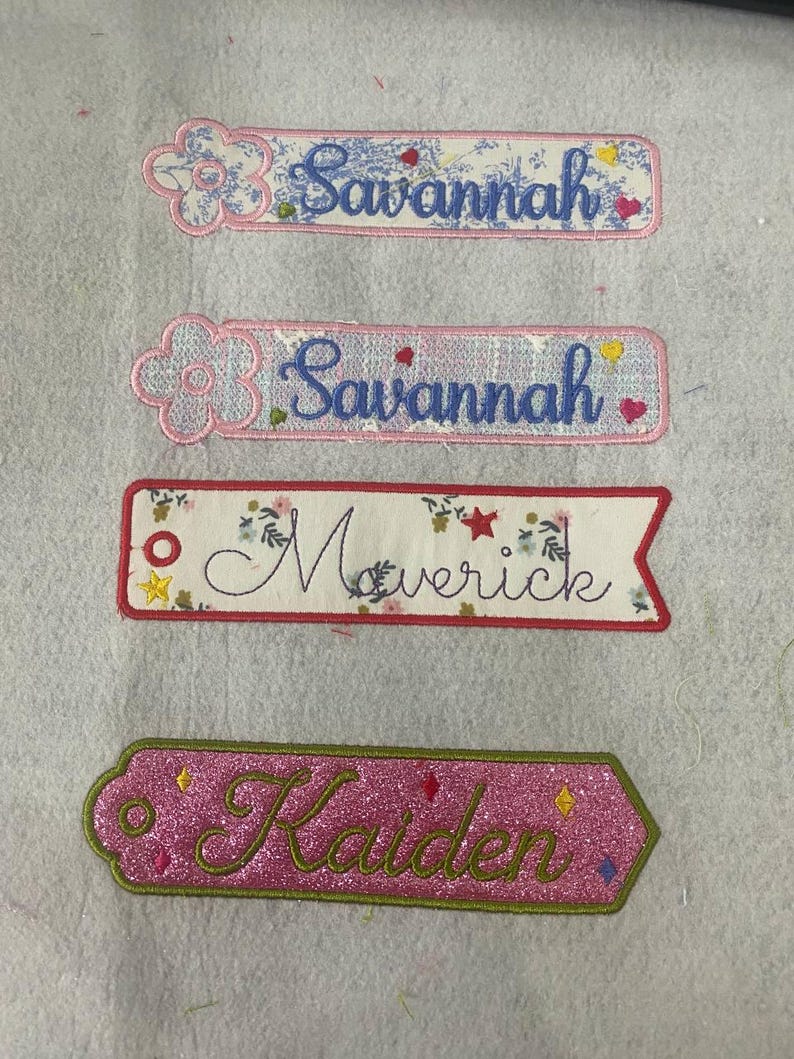May include: Four embroidered fabric name tags. Two rectangular tags feature the name Savannah in blue script with a flower icon. One rectangular tag with a pointed end features the name Maverick in black script with floral and star accents. One pink glitter tag features the name Kaiden in green script.
