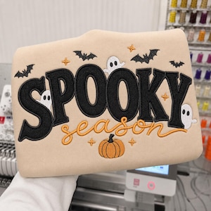 Könnte beinhalten: Ein beigefarbenes Sweatshirt mit schwarzer Stickerei des Wortes SPOOKY und orangefarbenem Schreibschrift-Text season. Das Design zeigt weiße Geisterfiguren, schwarze Fledermäuse, einen orangefarbenen Kürbis und kleine orangefarbene Sternsymbole.