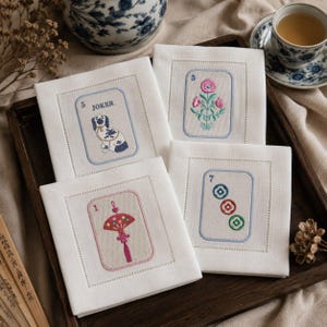 May include: Four white linen coasters with embroidered card-themed designs. Each coaster features a different design: a dog, flowers, a fan, and colorful circles. The coasters are arranged on a wooden tray, with a teacup and decorative vase in the background.