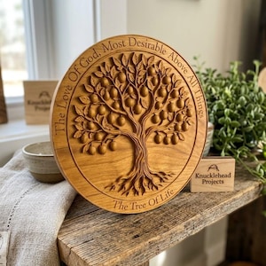 May include: A circular wooden plaque featuring a relief carving of a tree with fruit and roots. The text "The Love Of God, Most Desirable Above All Things" is engraved along the top edge, and "The Tree Of Life" is engraved along the bottom edge.