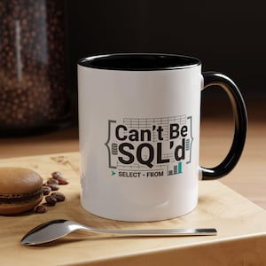 May include: White ceramic mug with a black interior and handle. The mug features the text "Can't Be SQL'd" in a gray and teal design. A spoon, a macaron, and coffee beans are on a wooden surface.