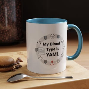 May include: White ceramic mug with a blue interior and handle. The mug features the text "My Blood Type Is YAML" surrounded by computer-related icons. A spoon and a pastry are in the foreground.