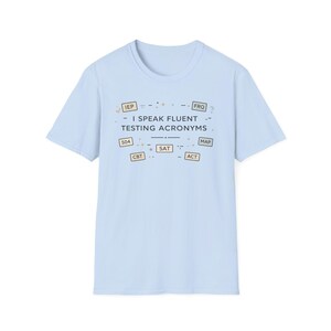 May include: Light blue t-shirt with the text "I SPEAK FLUENT TESTING ACRONYMS." The shirt features various acronyms such as IEP, FRQ, 504, CBT, SAT, MAP, and ACT in small boxes. The shirt is made of a soft material.