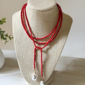 May include: A red beaded necklace with a lariat design. The necklace features two strands of small red beads that meet at a knot, with two white, irregular-shaped pearl pendants hanging from the ends. The necklace is displayed on a mannequin.