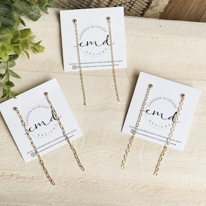 May include: Gold-colored chain earrings displayed on white cards with the text "Cameron McIntosh Designs." The earrings have a small stud at the top and a long chain that dangles down. The cards also include a website and social media handle.