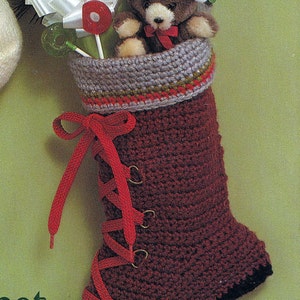 May include: A red crocheted stocking with a brown cuff and red laces. The stocking is filled with a small stuffed teddy bear wearing a red Santa hat, a white bow, and a red lollipop.