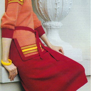 Puede incluir: Una mujer con un suéter a rayas rojas y naranjas, una falda roja y un sombrero rojo. Está de pie junto a una urna blanca.