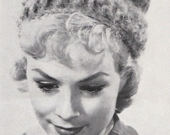May include: A black and white photo of a woman wearing a knitted hat with a textured design. The hat is worn low on her forehead and covers her ears. The woman is looking down and smiling.
