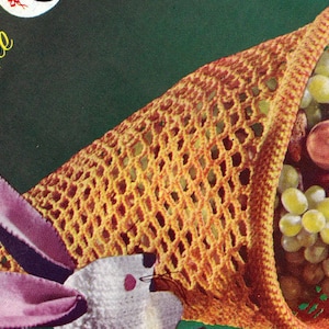 Puede incluir: Una cornucopia de crochet naranja y amarilla llena de uvas verdes y nueces marrones. Un conejo rosa y blanco asoma por detrás de la cornucopia.