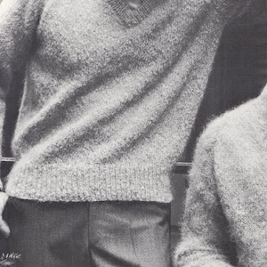 以下が含まれることがあります： Vネックのふわふわのライトグレーのセーターを着た男性の白黒写真。彼は笑顔で腕を上げている。男性はグレーのパンツを履いている。