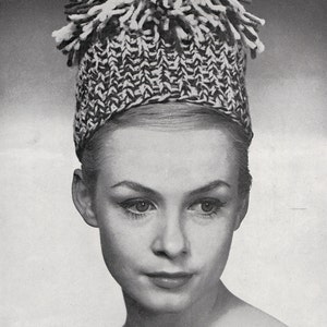 May include: A black and white knitted hat with a textured pattern and a fringe of yarn on top. The hat is worn by a person with fair skin and dark eye makeup. The image is in black and white.
