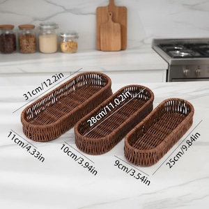 May include: Three brown woven wicker baskets of varying sizes. The largest is 12.2 inches long, 4.33 inches wide, and the smallest is 9.84 inches long and 3.54 inches wide. The baskets are empty and sit on a white countertop.