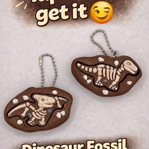 Peut inclure: Deux clickers fossiles de dinosaures bruns avec des d&eacute;tails d'os blancs et des porte-cl&eacute;s &agrave; cha&icirc;ne argent&eacute;e. Le texte "Tap -> You'll get it" est en haut et "Dinosaur Fossil Clickers" en bas.
