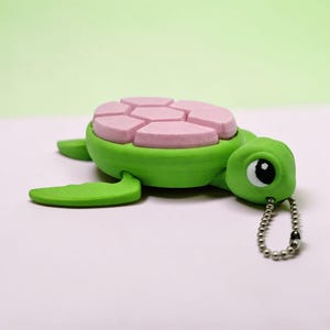 May include: A green turtle-shaped keychain with a pink segmented shell. The turtle has a detailed head with a large eye and a silver ball chain. The design is whimsical and playful, perfect for a gift or personal accessory.
