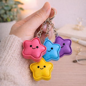 May include: A set of four star-shaped keychains in pink, blue, purple, and yellow. Each star features a simple black printed face with two circular eyes and a curved line for a mouth. The stars are attached to a silver metal ring by chains.