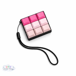 Könnte beinhalten: Ein quadratisches, schwarzes Gerät mit neun rosa und weißen Tasten in einem 3x3-Raster. Ein schwarzes Schlüsselband ist befestigt. Die Tasten sind in verschiedenen Rosatönen gehalten. Das Gerät befindet sich auf weißem Hintergrund.
