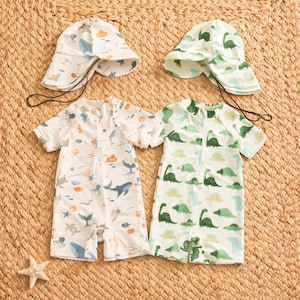 Puede incluir: Dos trajes de baño para bebés y dos sombreros para el sol a juego. Un traje de baño es blanco con un estampado de vida marina azul, naranja y gris. El otro es blanco con gráficos de dinosaurios verdes. Los sombreros tienen patrones a juego y correas para la barbilla.