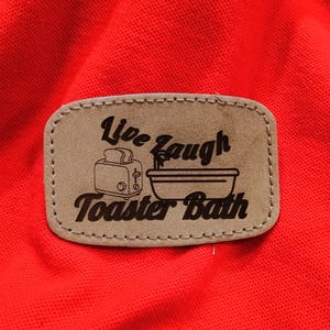 May include: A brown leather patch with the words "Live Laugh Toaster Bath" in a whimsical font. The patch features a toaster and a bathtub graphic. The patch is sewn onto a bright red fabric.