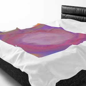 May include: A soft throw blanket featuring a vibrant gradient pattern of purple, pink, and orange hues. The blanket is draped over a white bed sheet, with the fabric showing gentle folds and a smooth texture across the colorful surface area.