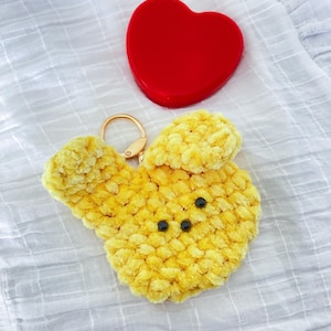 May include: A yellow, plush bunny keychain with black bead eyes and a gold-colored keyring. A red heart-shaped object is next to the keychain. The items are on a white, textured surface.