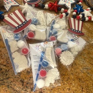 May include: A set of patriotic craft kits in clear plastic bags, each containing white ceramic shapes, a small paintbrush, and three small paint pots in red, white, and blue. The kits feature labels with text including 4th of July and instructions.