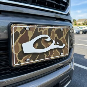 Puede incluir: Una placa de matr&iacute;cula rectangular con un patr&oacute;n de camuflaje en tonos marr&oacute;n, beige y negro. Un logotipo grande y blanco de una cabeza de pato estilizada est&aacute; centrado en la placa. El marco est&aacute; sujeto al parachoques gris con cuatro tornillos.