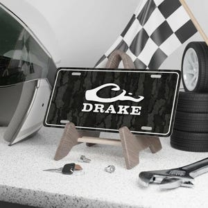 May include: A black and white license plate with the name "DRAKE" in white lettering, displayed on a wooden stand. The plate has a camouflage pattern. A checkered flag, tires, and a helmet are in the background.