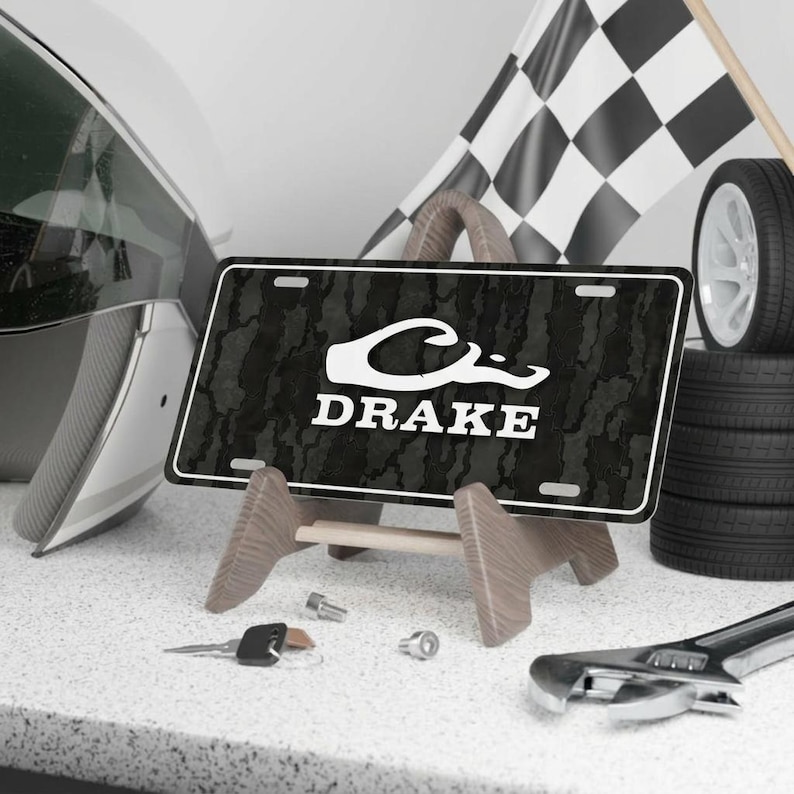 May include: A black and white license plate with the name "DRAKE" in white lettering, displayed on a wooden stand. The plate has a camouflage pattern. A checkered flag, tires, and a helmet are in the background.