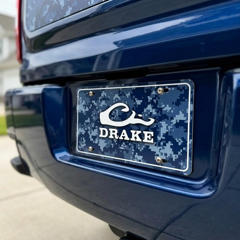 May include: A blue license plate with a camouflage pattern and the name "DRAKE" in white lettering. The plate is mounted on a dark blue vehicle bumper. The plate features a stylized wave design above the name.
