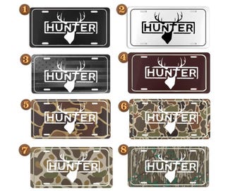 May include: Collection of eight license plates with the word "HUNTER" and a deer silhouette. The plates feature various designs, including black, white, camouflage, and an American flag.