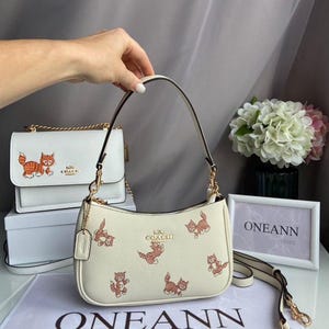 May include: Cream-colored Coach handbag with a repeating pattern of orange and brown cartoon cats. The bag has a curved shape, a top handle, and a detachable shoulder strap. A smaller matching crossbody bag is in the background.