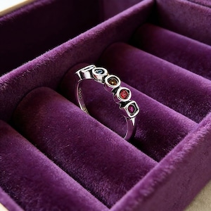 May include: A silver ring with five colorful gemstones, including blue, green, and red, set in a row. The ring is displayed in a purple velvet jewelry box, showcasing its design and craftsmanship. The gemstones are square and round.