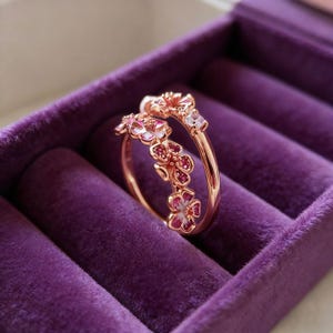 May include: A rose gold ring featuring a floral design with pink and white enamel petals and small red accents. The ring is set against a purple velvet background, showcasing its delicate details and craftsmanship.