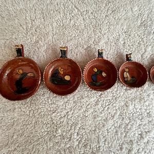 May include: A set of five handmade, terracotta-colored ceramic bowls with handles. Each bowl features a unique hand-painted scene in black, yellow, and blue. The bowls vary in size, with the largest being approximately 6 inches in diameter.