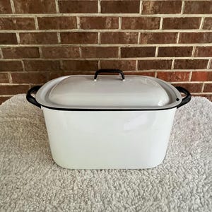 May include: White enamel rectangular baking dish with a lid. The dish has black trim and handles. The lid has a black handle. The dish is sitting on a white, textured surface in front of a brick wall.