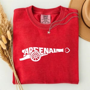 May include: A red t-shirt with the word "ARSENAL" in white, designed as a cannon. The shirt has a crew neck and a small tag. A silver necklace with a star pendant is draped over the shirt. A brown hat is visible in the background.