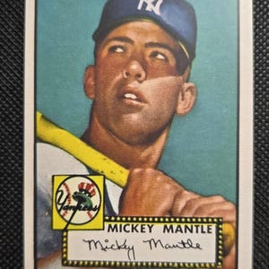 Puede incluir: Tarjeta de béisbol vintage con Mickey Mantle de los Yankees de Nueva York. La tarjeta muestra un retrato de Mantle con una gorra de los Yankees, sosteniendo un bate. El logo de los Yankees es visible. La tarjeta tiene un borde blanco y un fondo verde azulado.