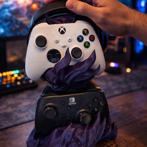 May include: A white Xbox controller and a black Nintendo Switch controller rest on a purple flame-like stand. A black headset sits above the Xbox controller. The controllers have various colored buttons.