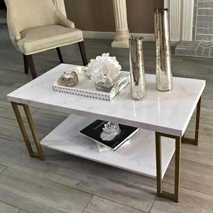 May include: A white marble-topped coffee table with gold-colored metal legs. The table has a lower shelf and is decorated with vases, a tray of decor, and a book. The table is in a living room setting.