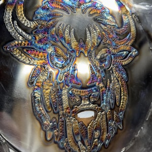 Puede incluir: Una obra de arte en metal que representa una cabeza de bisonte, creada con técnicas de soldadura. La pieza presenta un diseño detallado con una gama de colores, incluyendo azules, dorados y púrpuras, logrados mediante tratamiento térmico. El bisonte tiene grandes cuernos y una barba detallada.