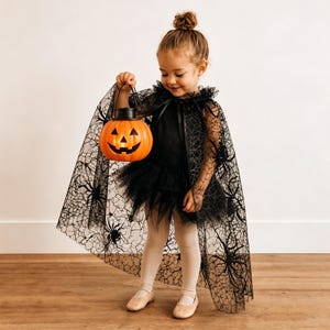 K&ouml;nnte beinhalten: Ein schwarzer, transparenter Umhang mit Spinnennetz- und Spinnenmuster &uuml;ber einem schwarzen Kleid mit T&uuml;llrock. Das Outfit wird mit hellen Leggings und Ballerinas kombiniert, w&auml;hrend ein orangefarbener K&uuml;rbis-Eimer aus Kunststoff gehalten wird.